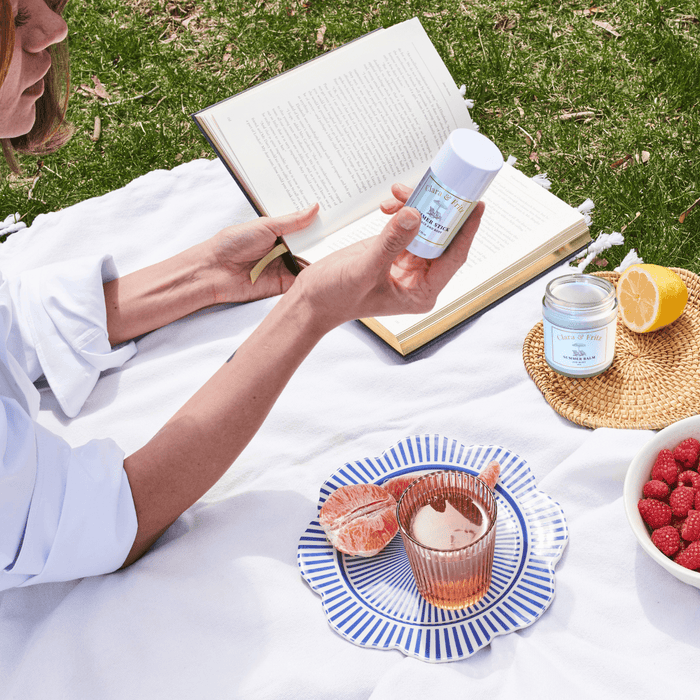 Woman enjoying a picnic with Sun-kissed Trio products, including a skincare bottle and candle, surrounded by fresh fruit and an open book.