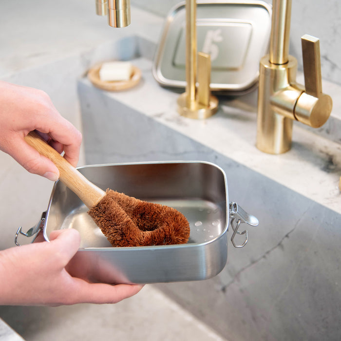 Hand holding a wooden dish brush with brown bristles over a metal container on a kitchen counter.