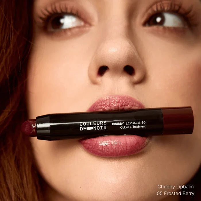Close-up of a woman's lips with a chubby lipbalm labeled 'Couleurs De Noir' held between her lips.