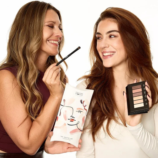 Two women holding makeup products and smiling against a white background